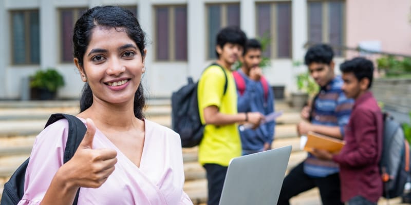 A confidant girl student showing thumbs up at college campus while using laptop.